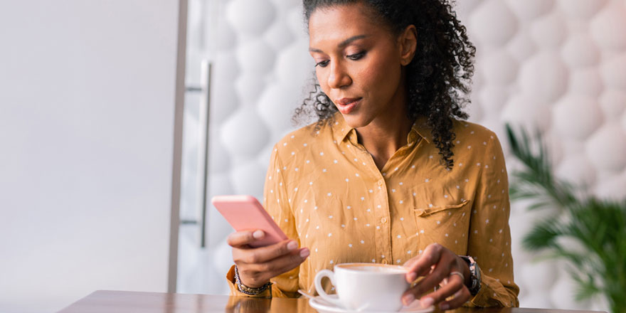 A young woman on her phone is drinking coffee and looking at her Premier Money Market account online