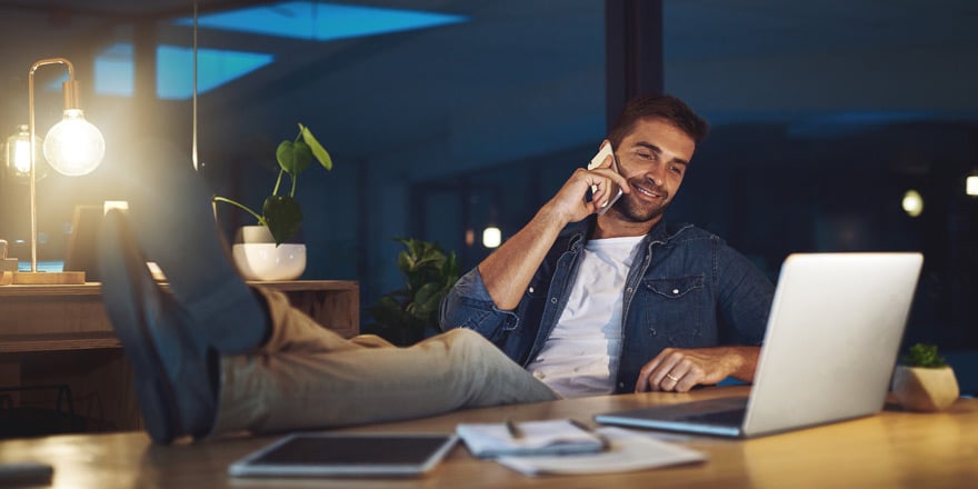 A younger man relaxes at his office desk and uses his laptop and mobile device for telephone banking