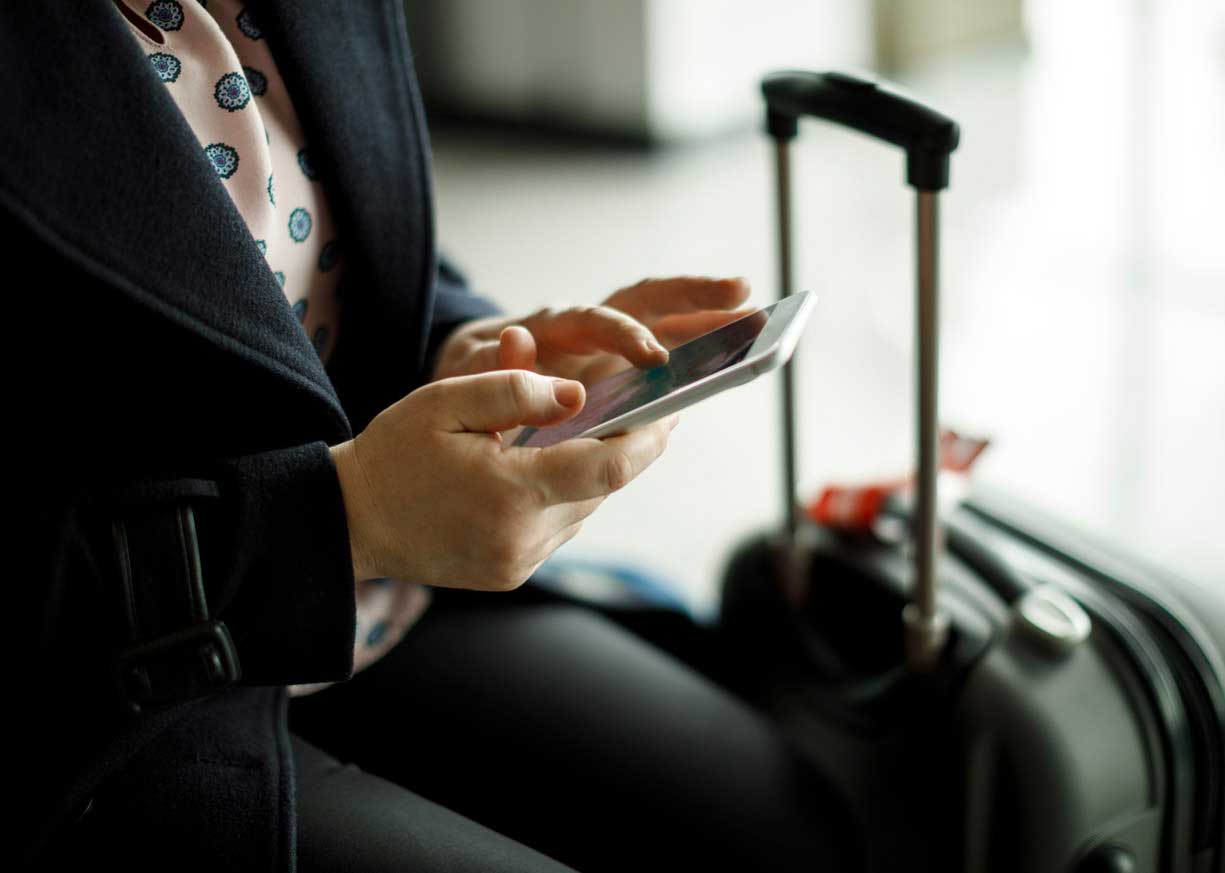 A business professional traveling with a suitcase in an airport compares commercial checking accounts on a mobile device