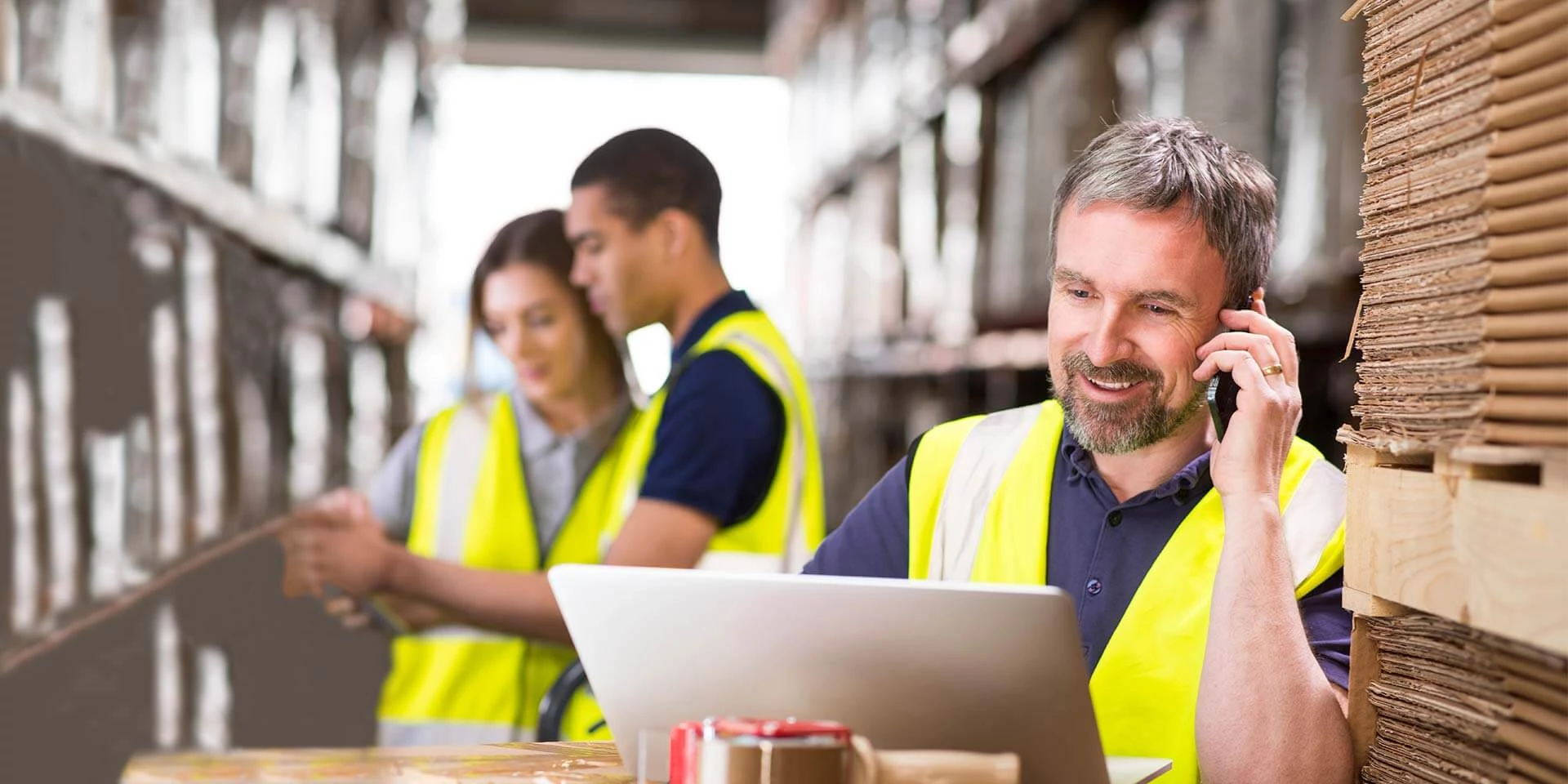 A business manager in a shipping warehouse is using his phone and laptop to access online banking and the banking app